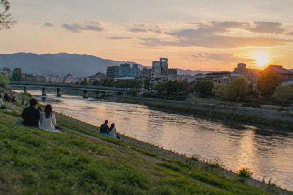 People sitting on the grassy banks of the Kamo river at dusk with the city skyline behind