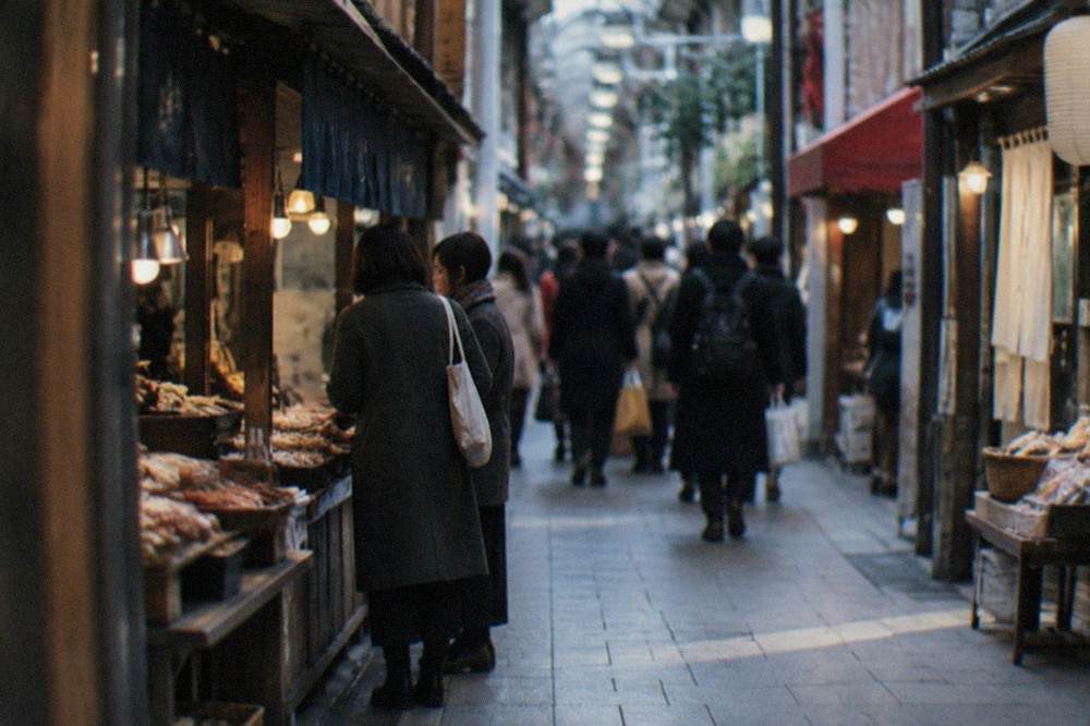 A narrow covered market lane in Kyoto lined with food stalls and shoppers under ceiling lamps