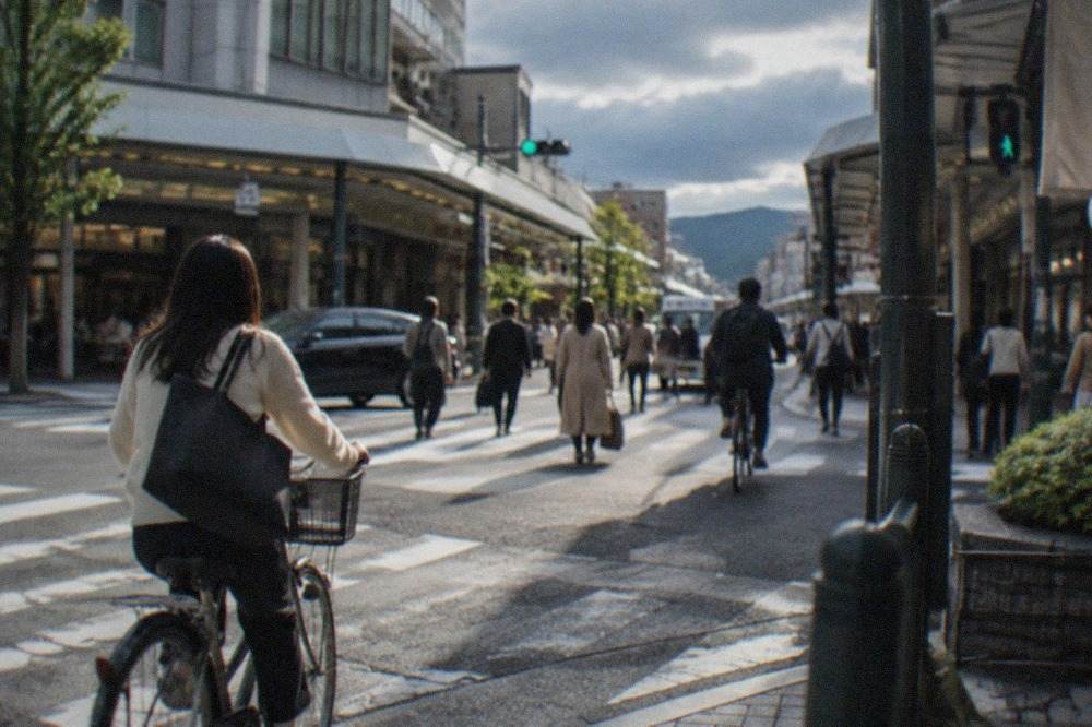 Shoppers and cyclists crossing a busy downtown Kyoto intersection lined with department stores and narrow side streets