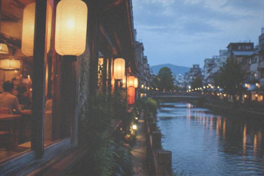 A narrow lantern-lit alley beside a canal in downtown Kyoto at dusk, with diners visible through restaurant windows