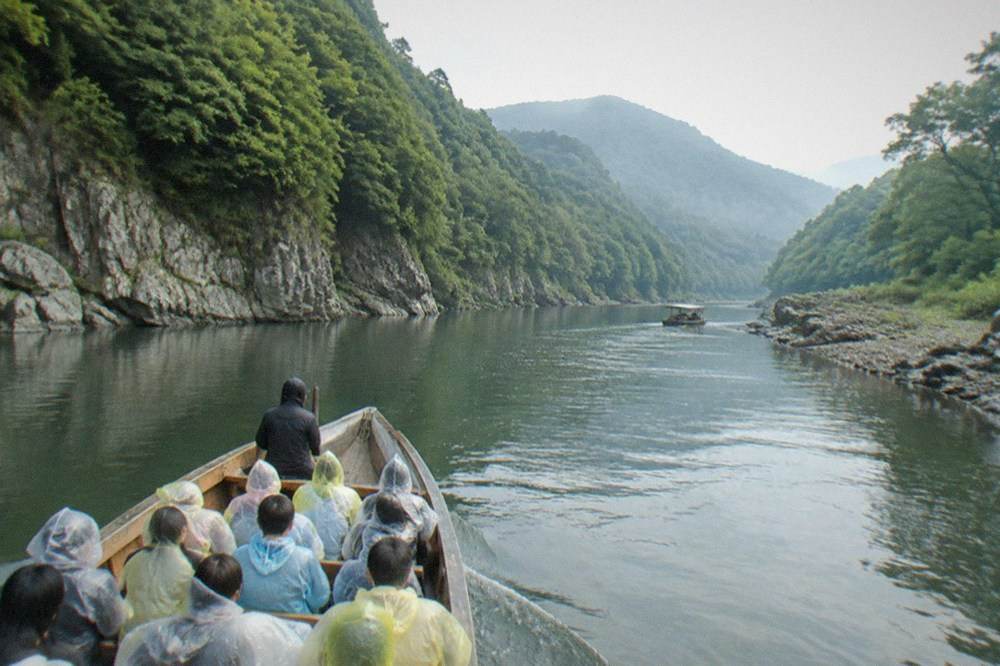 Passengers in rain ponchos on a wooden boat moving through calm water between green cliffs