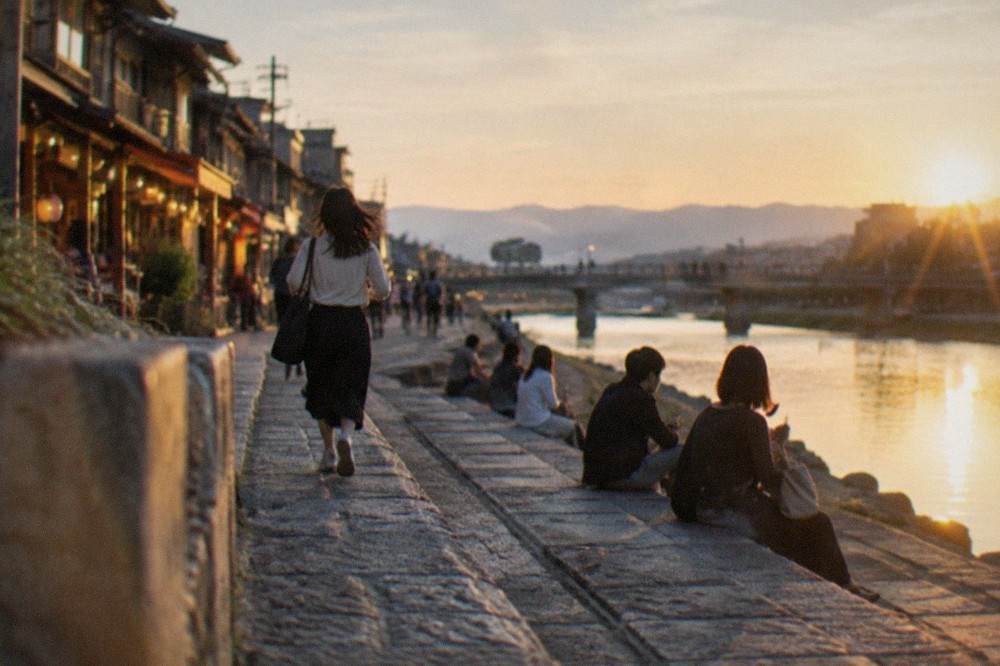 People sitting on wide stone steps beside a river at dusk with lit shopfronts behind