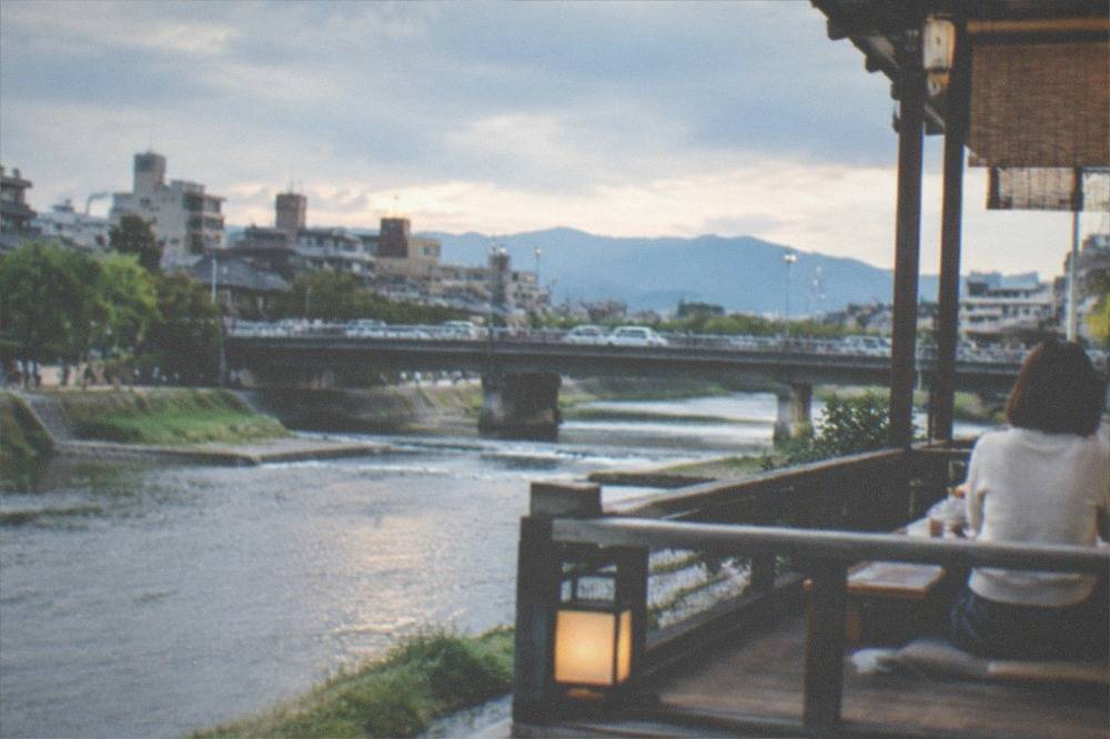 A diner seated at a low table on a wooden platform above a river in the early evening