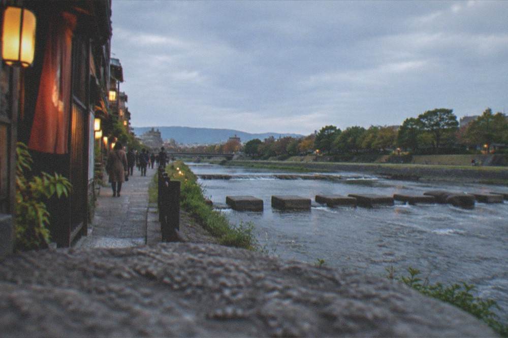 A wide river with stepping stones crossing it and a tree-lined bank at dusk