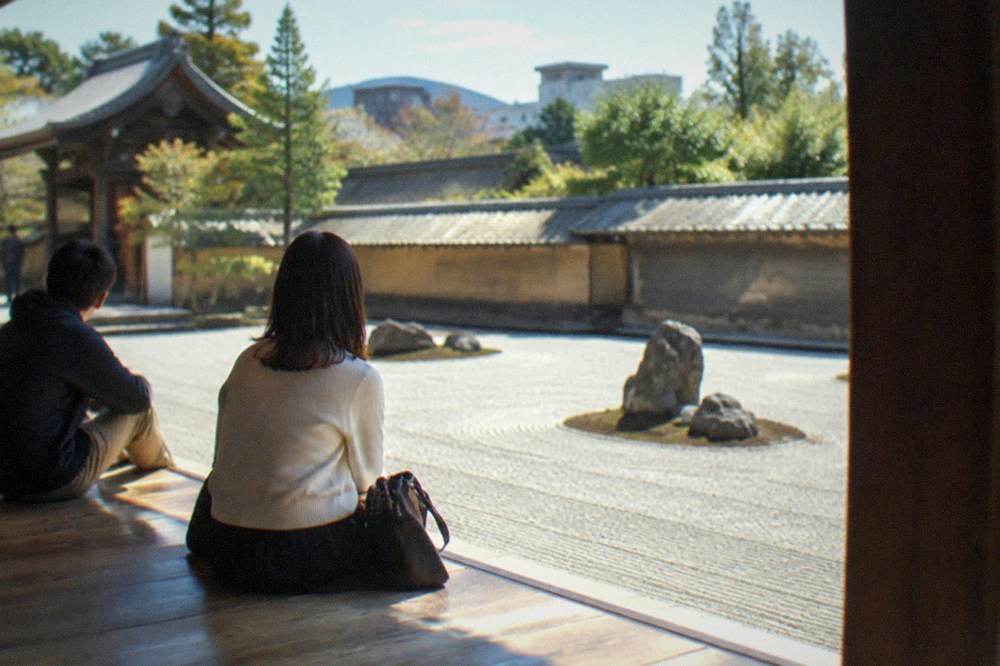 Visitors seated on a wooden temple veranda looking out over a rectangular rock garden of raked gravel and stones