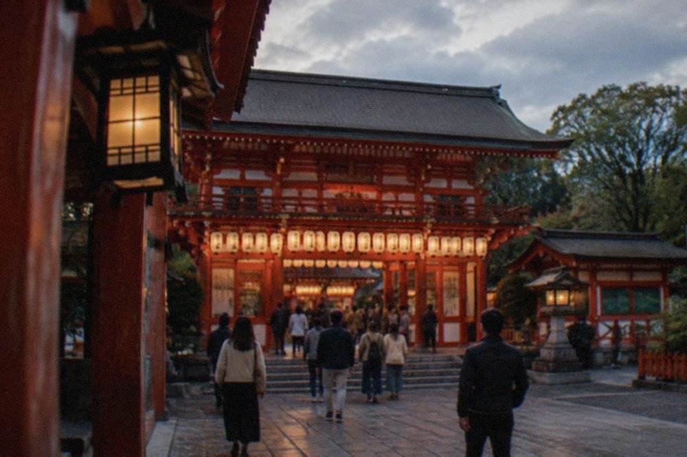 The lantern-lit main gate of Yasaka Shrine in Gion, Kyoto, with visitors walking through in the early evening