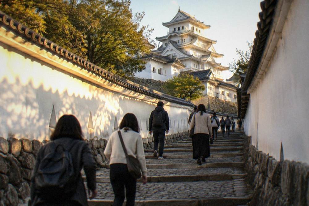 Visitors walking up a steep stone ramp between white plaster walls inside the castle complex