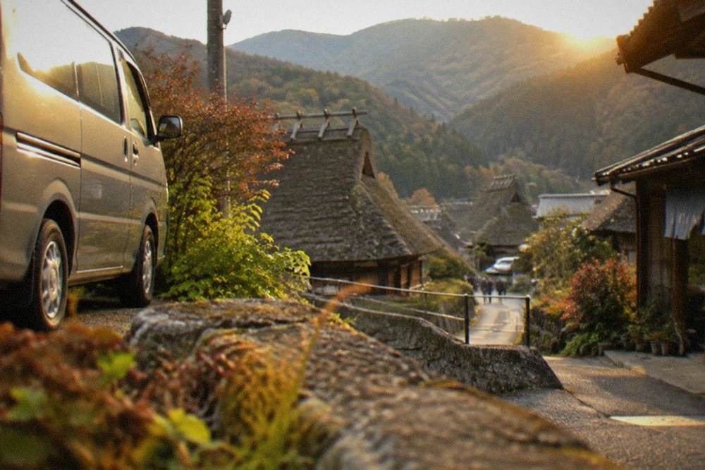 A private van parked on a narrow mountain road above a village of thatched houses