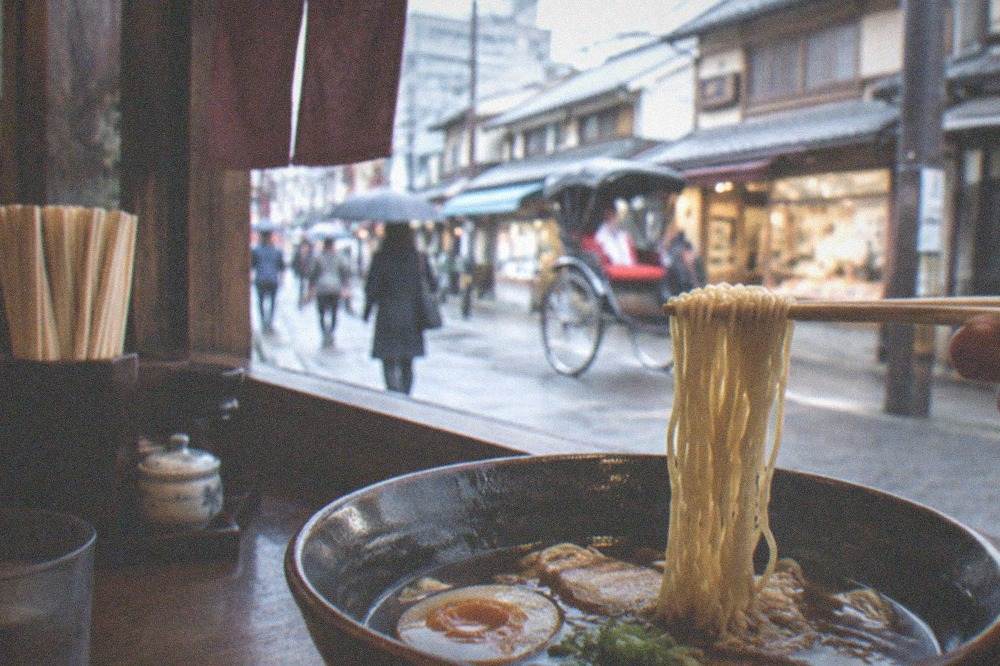 A bowl of dark Kyoto-style ramen with noodles being lifted by chopsticks at a counter