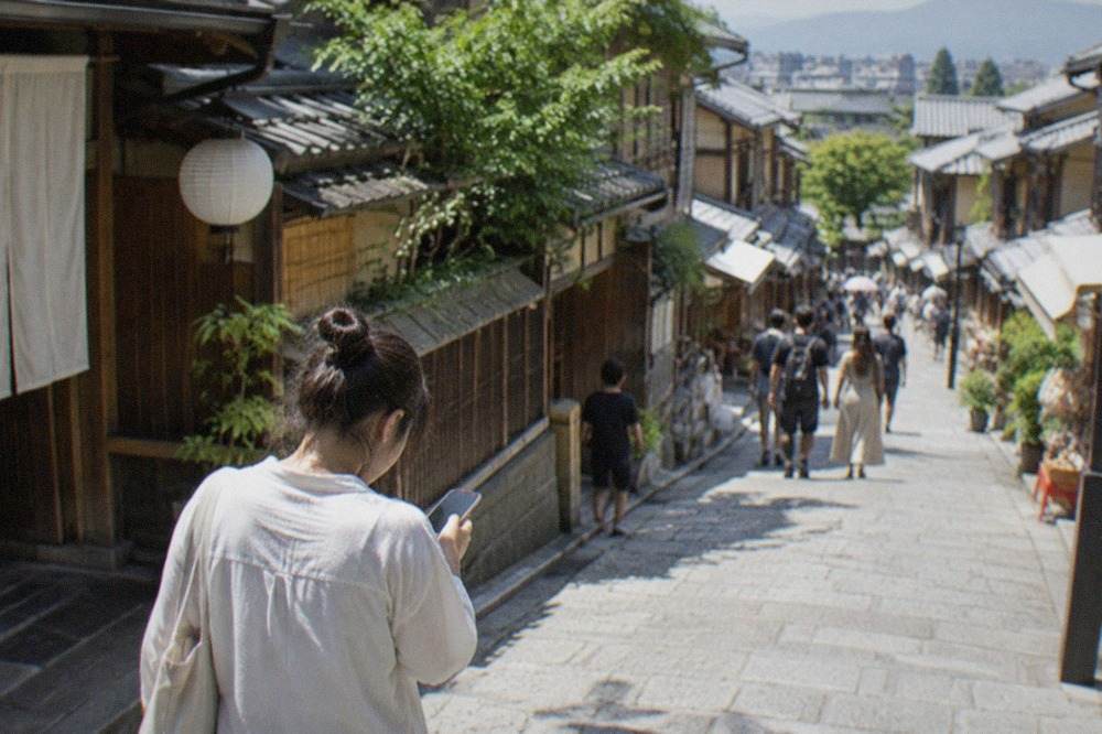 A traveller checking a phone on a stone street lined with traditional wooden machiya buildings in Kyoto