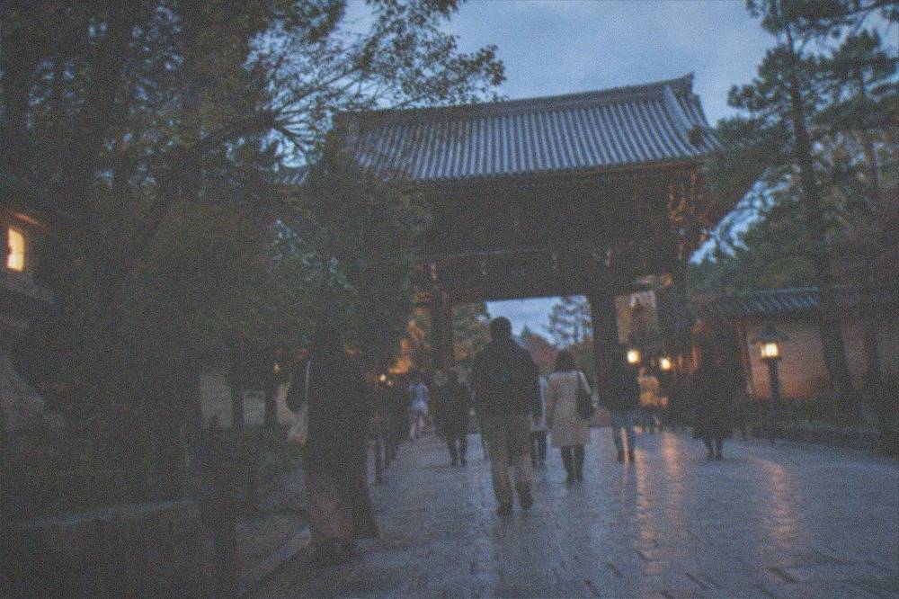 Visitors walking the stone approach path toward a large wooden temple gate surrounded by trees