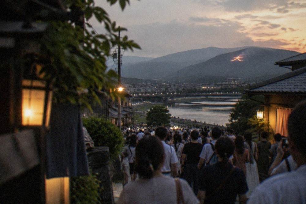 Crowds gathered on a Kyoto street at night watching distant bonfires glow on the mountains