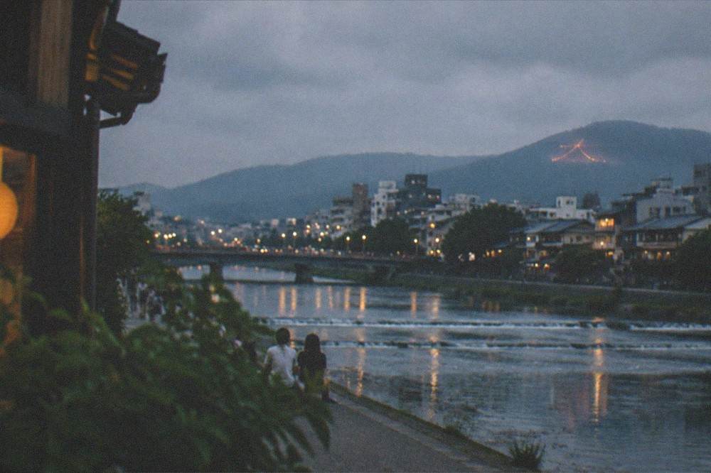 The Kyoto city skyline at night with a faint fire shape still glowing on a distant mountain