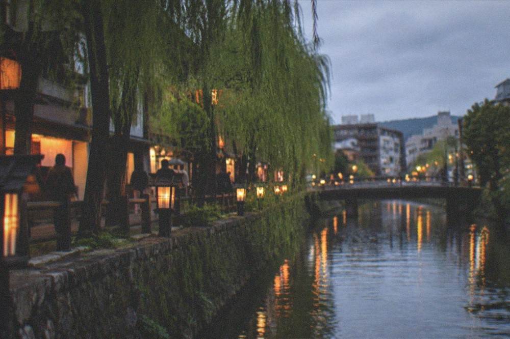 An evening view of a Kyoto canal with willow trees and warm lantern light