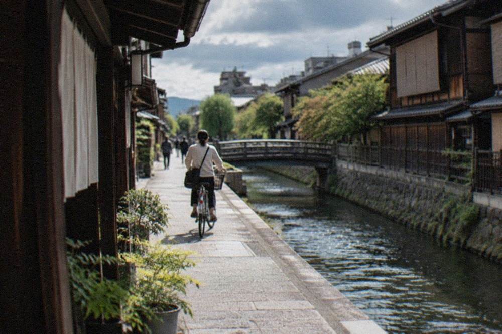 A cyclist riding along a quiet canal-side street in Kyoto with traditional wooden machiya houses on both sides