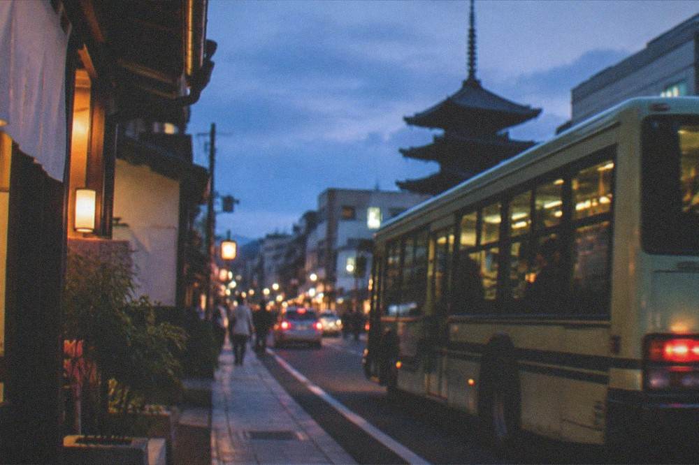 A Kyoto street at dusk with a bus passing illuminated shopfronts and a pagoda silhouette