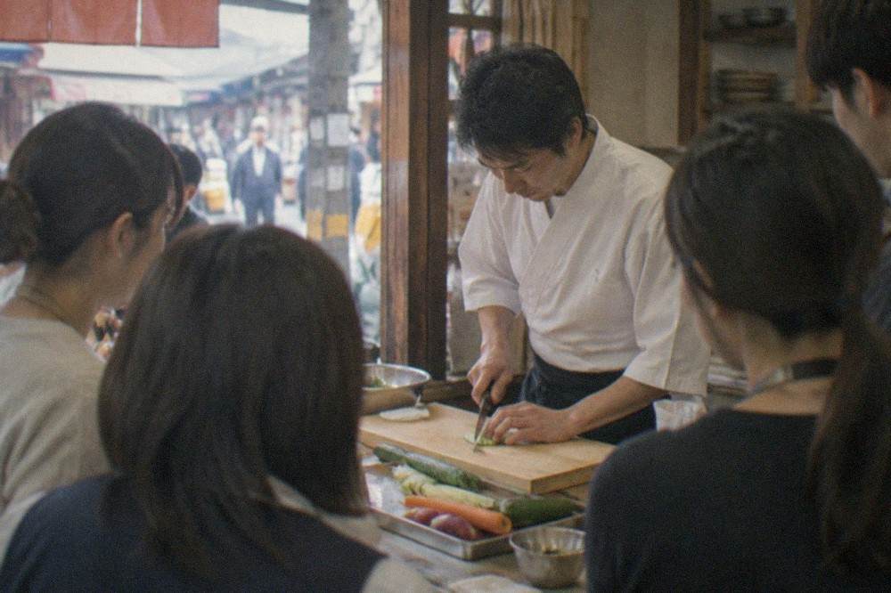 A instructor demonstrating vegetable knife work to students around a kitchen counter in Kyoto