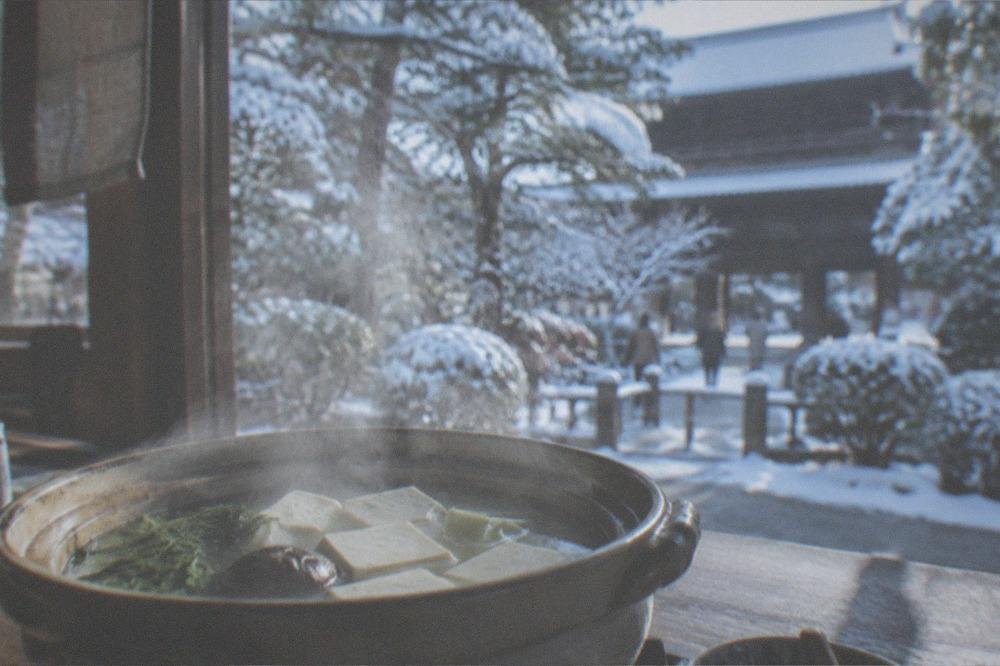 A steaming donabe pot of yudofu on a table beside a snowy temple garden window